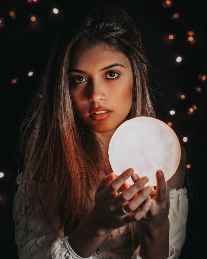 Portrait of a woman illuminated by a glowing sphere in a dark, fairy-lit background.